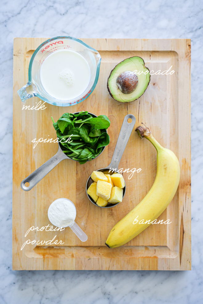 Avocado smoothie ingredients on a wooden cutting board with ingredient labels written in white letters.