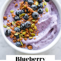 Bowl of blueberry smoothie topped with blueberries, granola, and pumpkin seeds with the words "Blueberry Smoothie Bowl" written in black underneath and "Fed + Fit" in golden orange.