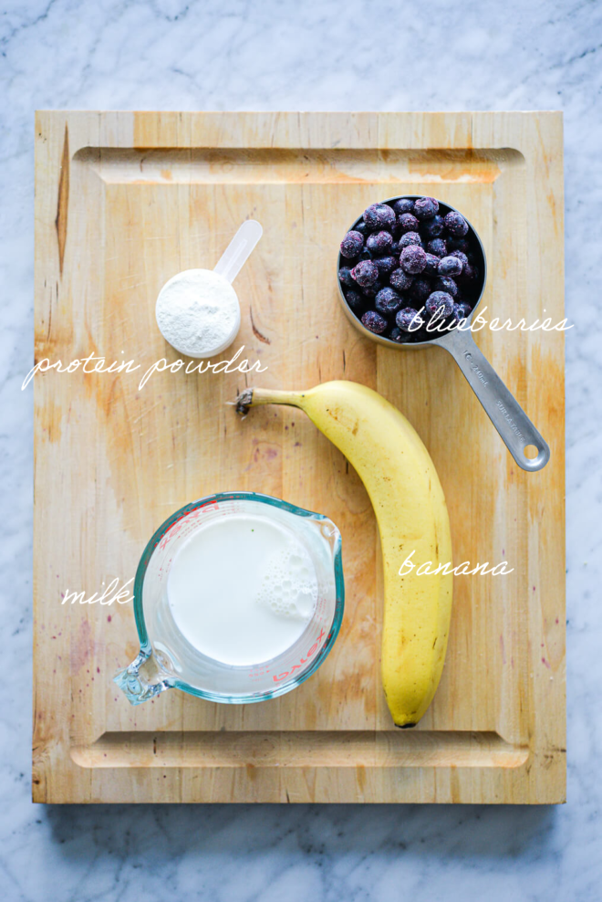 Blueberry smoothie ingredients on a wooden cutting board with ingredient labels written in white letters.