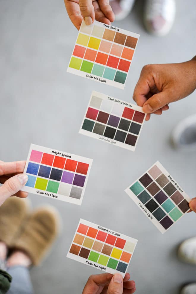 Top down view of hands holding 5 color template cards labeled: True Spring, Cool Sultry Winter, Brown Summer, Bright Spring, and Vibrant Autumn. All with Color Me Light on the bottom.