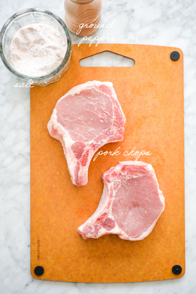 Two, raw bone-in pork chops on an epicurean cutting board with a jar of sea salt and pepper grinder on a marble surface.