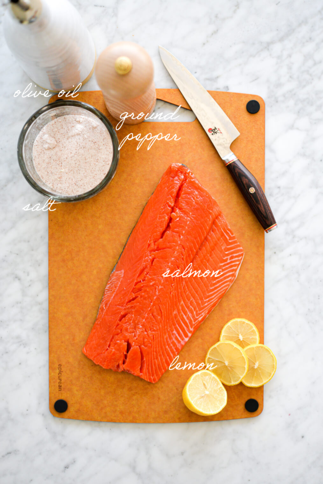 Raw salmon filet on an epicurean cutting board with sliced lemon, knife, a jar of sea salt, pepper grinder, and olive oil dispenser on a marble surface.