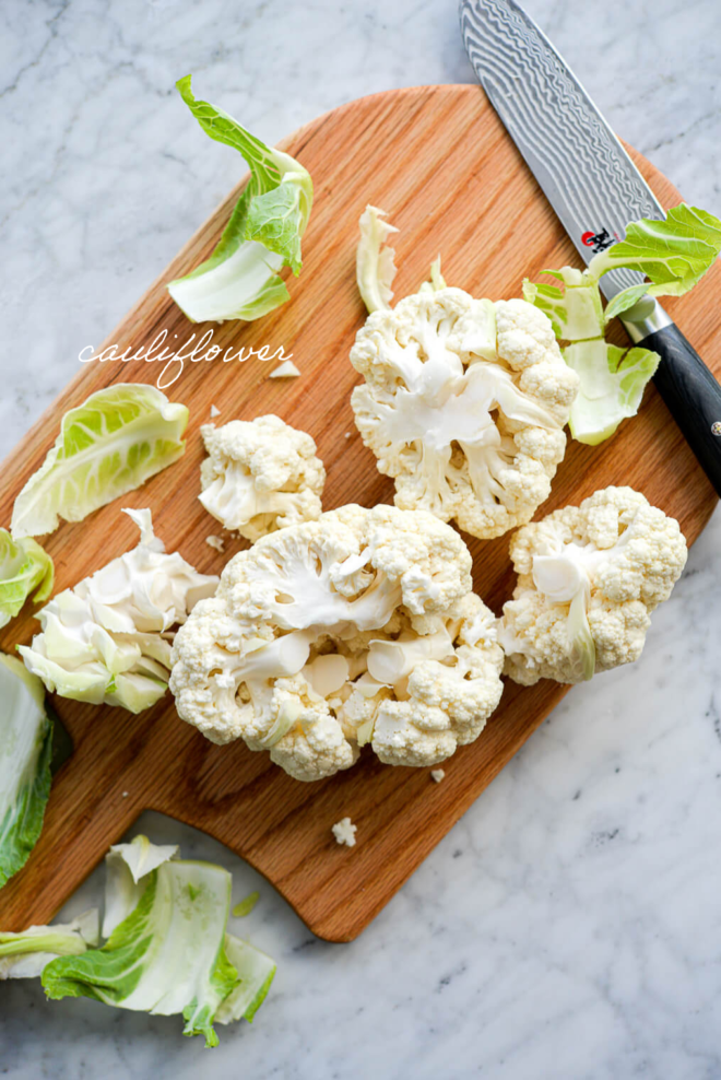 Cutting board with cauliflower florets ingredients on a marble surface with ingredient labels written in white letters.