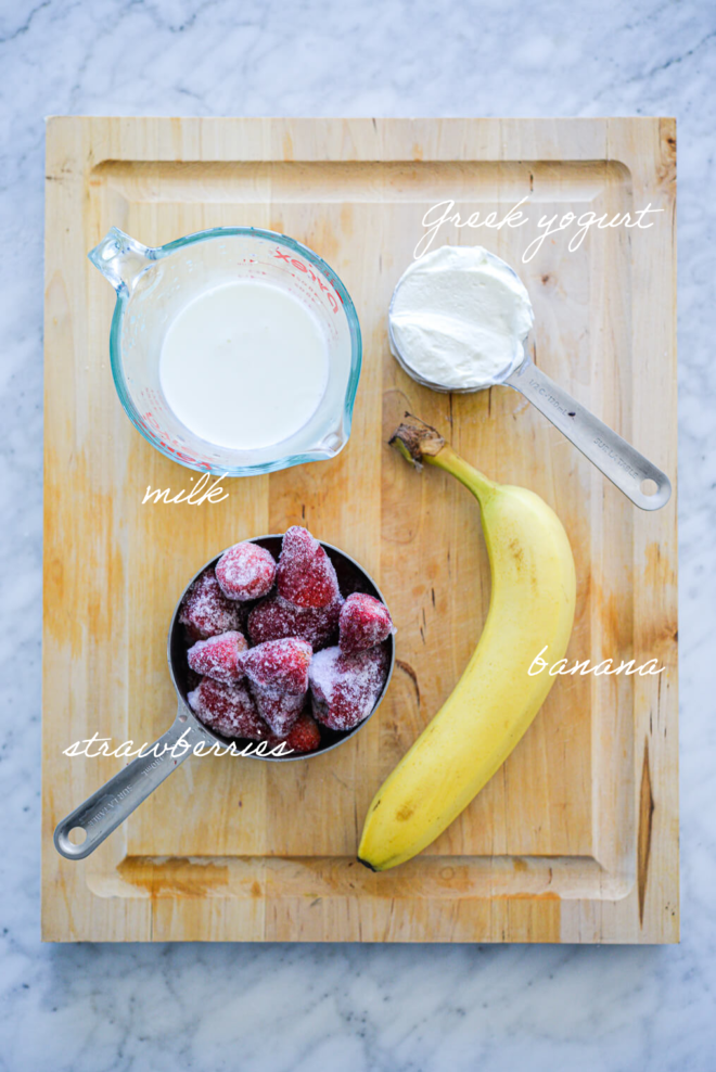 Strawberry smoothie ingredients on a wooden cutting board with ingredient labels written in white letters.