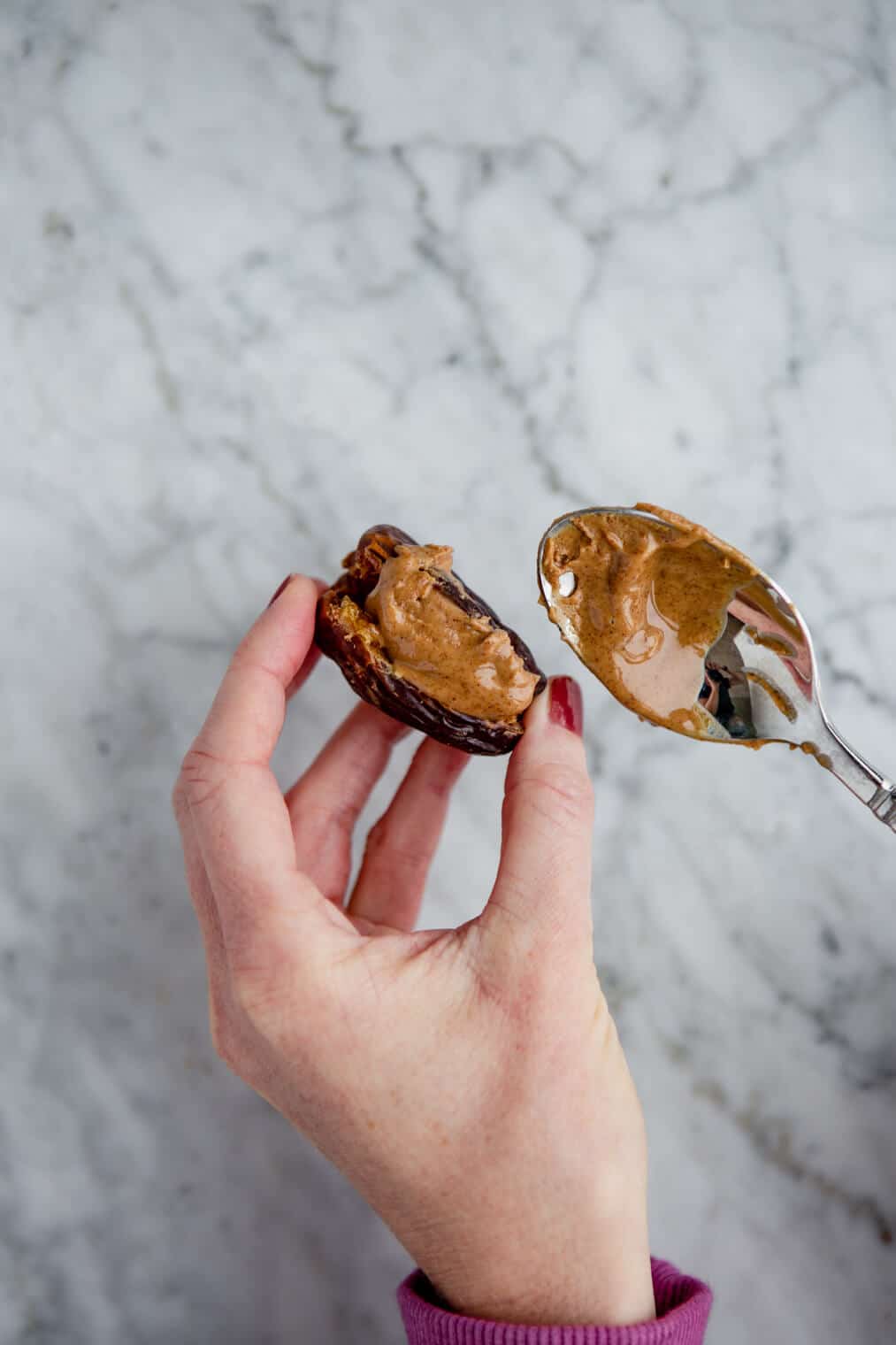 One hand holding pitted date filled with almond butter, other hand holding a spoon with almond butter.