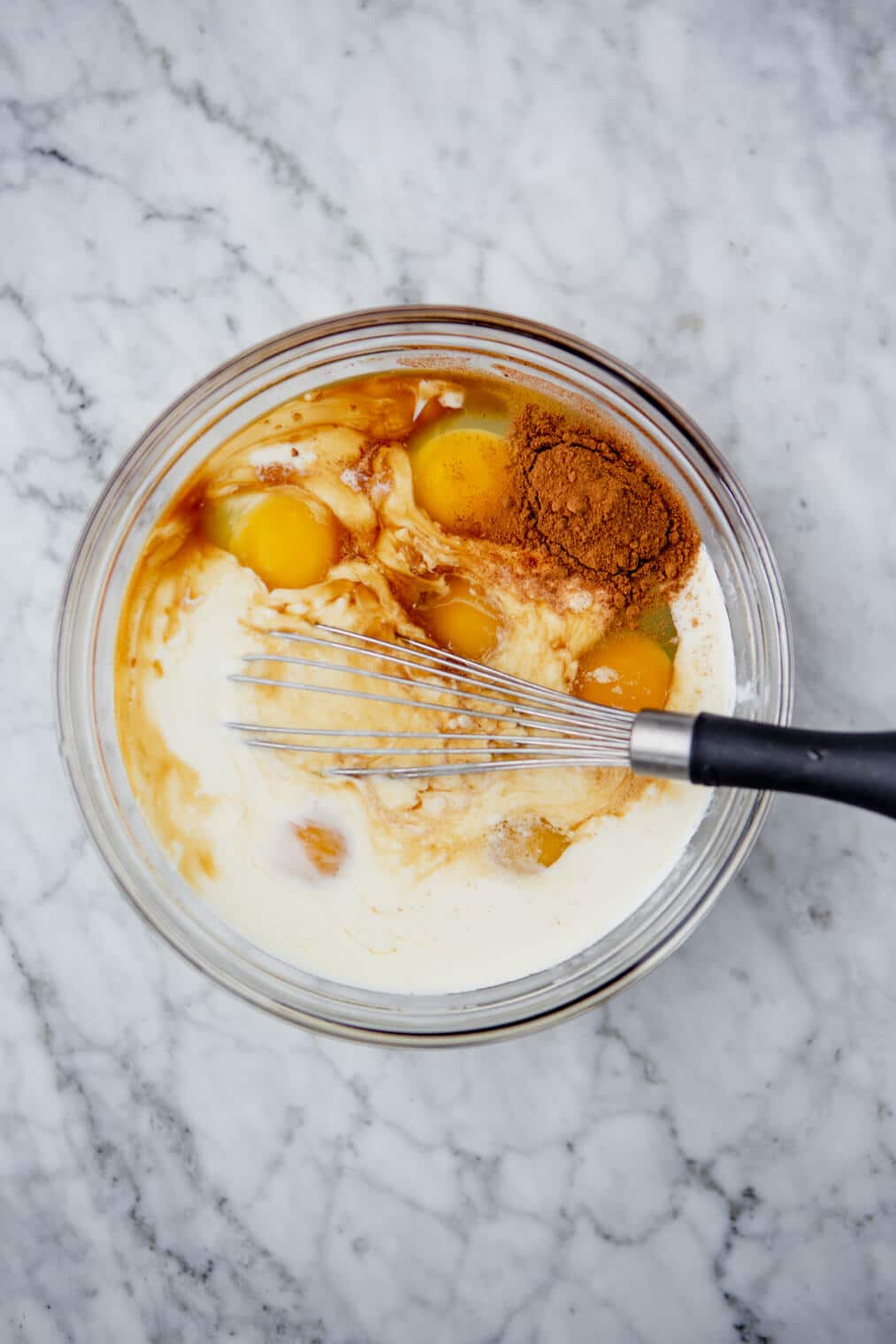 Bowl of eggs, milk, cinnamon, and vanilla with a whisk.