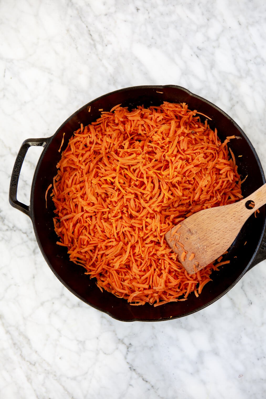 Cast iron skillet with shredded carrots evenly spread on bottom and wooden spatula.
