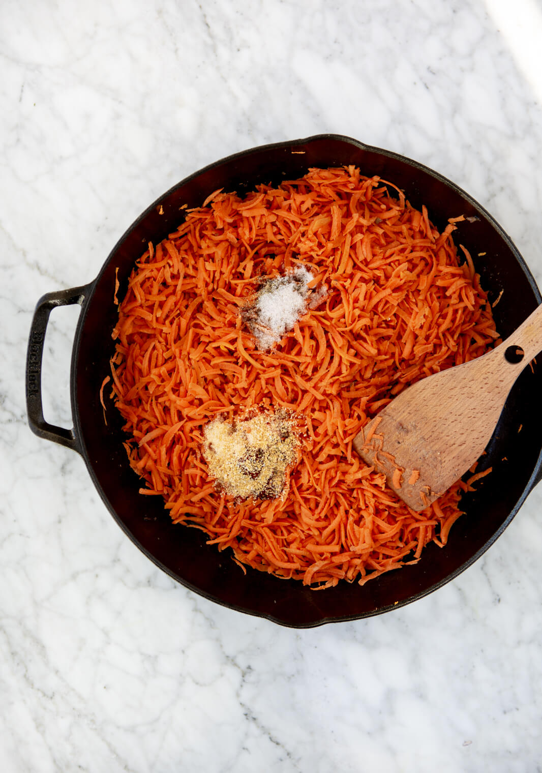 Cast iron skillet with shredded carrots evenly spread on bottom topped with spices.