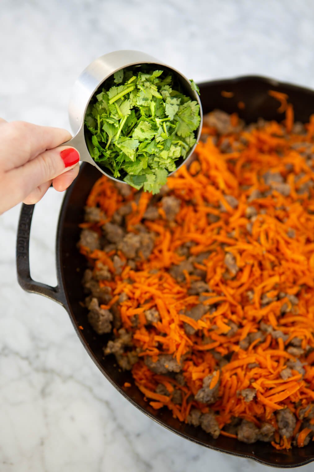 Measuring cup with chopped cilantro held over a cast iron skillet with shredded carrots and breakfast sausage.