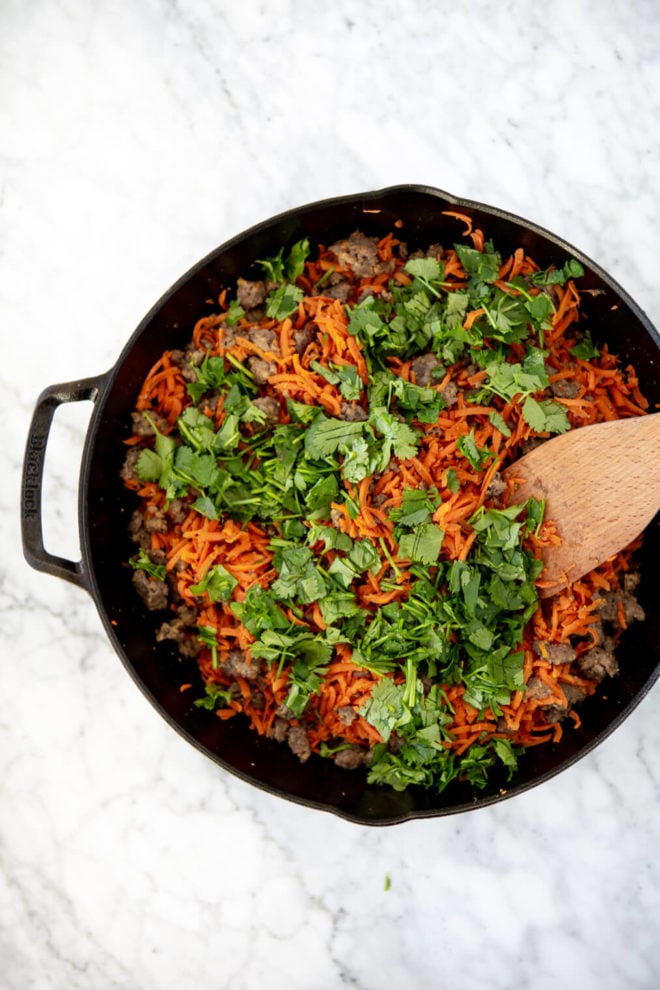 Cast iron skillet with shredded carrots, breakfast sausage, and chopped cilantro and wooden spatula.