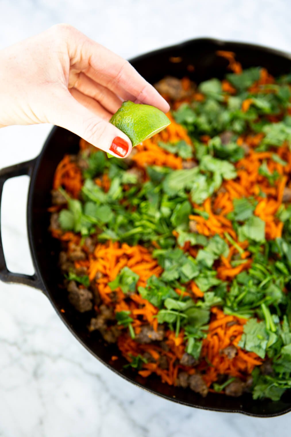 Wedge of lime being squeezed over cast iron skillet with shredded carrots, breakfast sausage, and chopped cilantro.