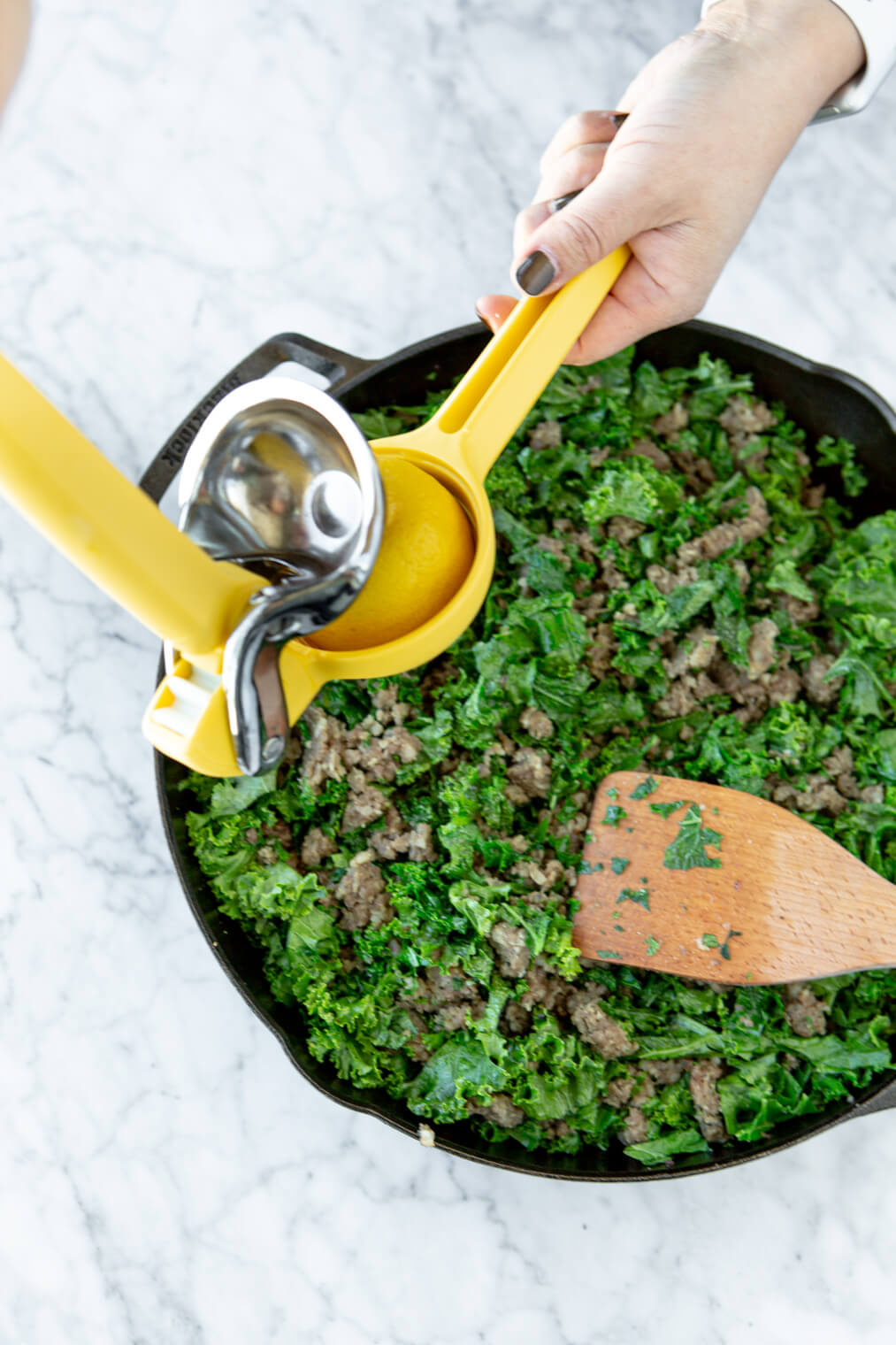 Lemon squeezer being held over the top of a cast iron skillet with chopped kale and sausage inside.