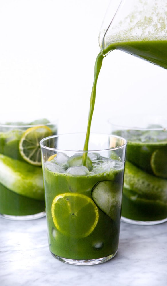 Three glasses lined with cucumber and lemon slices with ice and juice. Juice being poured into front glass from glass pitcher.