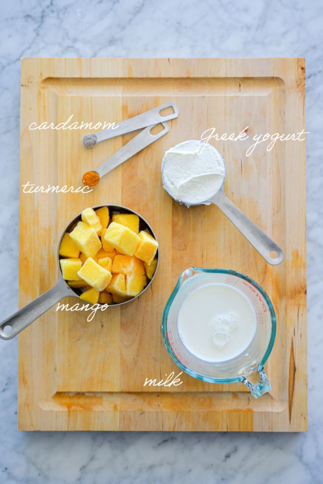 Mango smoothie bowl ingredients on a wooden cutting board with the name of the ingredients next to each item in white, cursive letters.