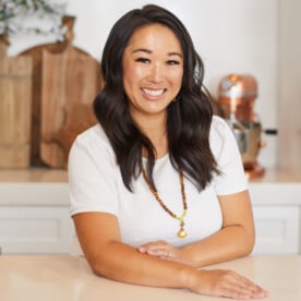 Waist up photo of Melissa wearing a cream colored shirt and mala bead necklace leaning on counter with one arm out in front and the other crossed in front of her.