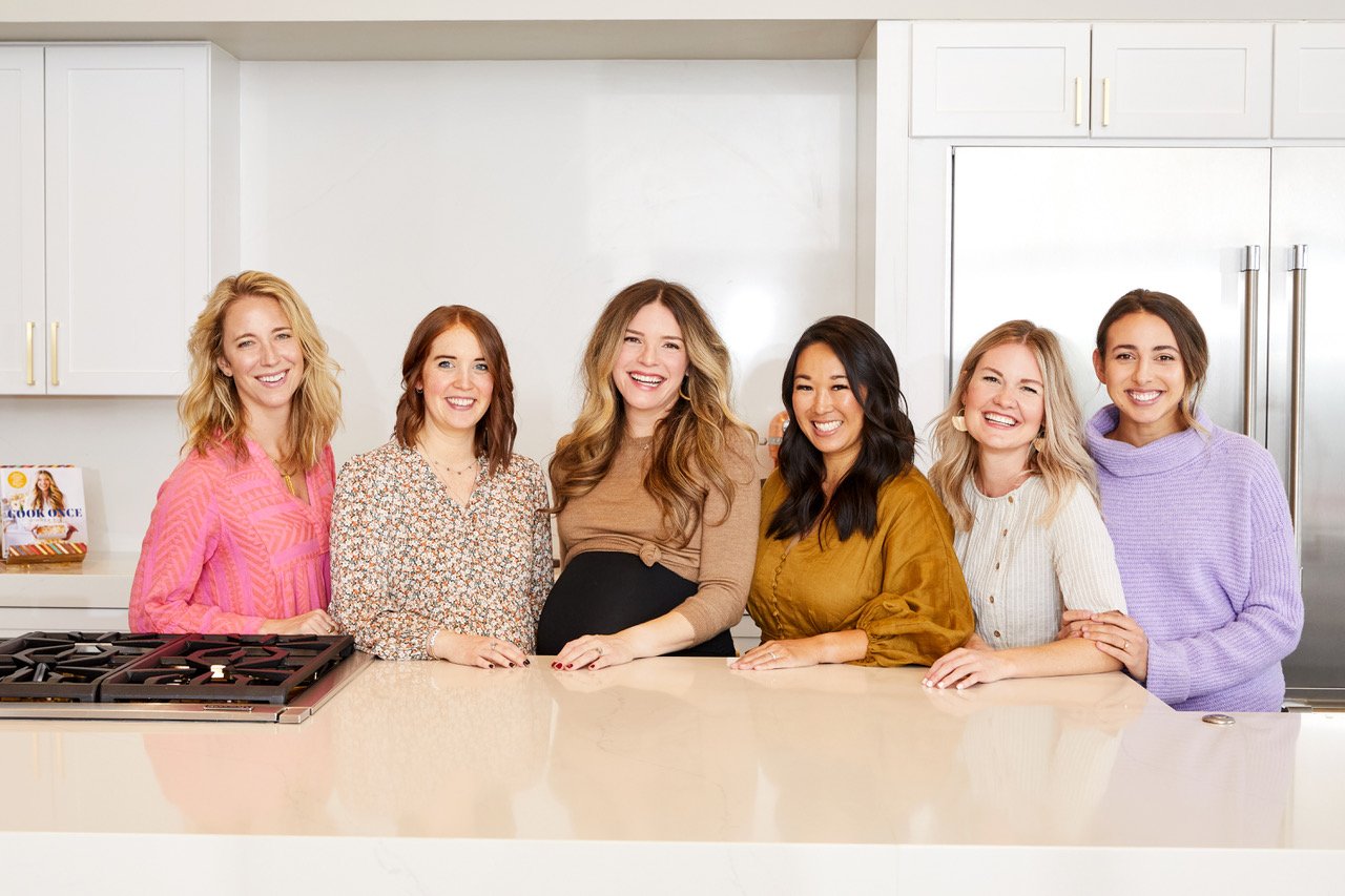 Fed + Fit Team smiling and standing at the kitchen counter (from left to right--Lindsay in pink long sleeved shirt, Brandi in teal and burnt orange floral shirt, Cassy in brown sweater and black dress, Melissa in mustard dress, Lauren in cream colored dress, and Graysen in lavender sweater).