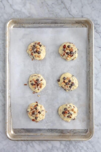 Cookie dough topped with additional toppings in three rows of two on a parchment lined baking sheet.