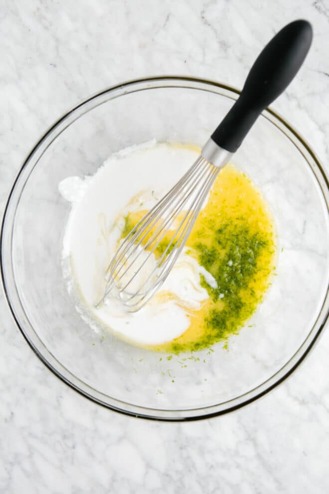 Egg, coconut milk, and lime zest in a glass mixing bowl on a grey and white marble surface.