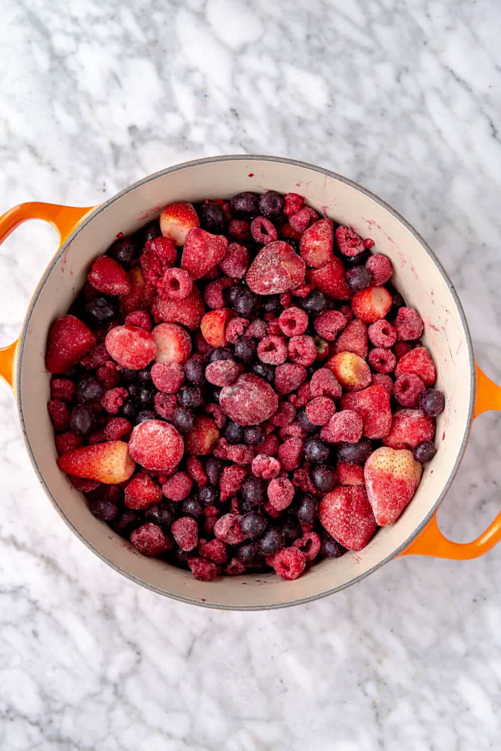 Frozen berries in a white enameled dutch oven with orange handles on a grey and white marble surface.
