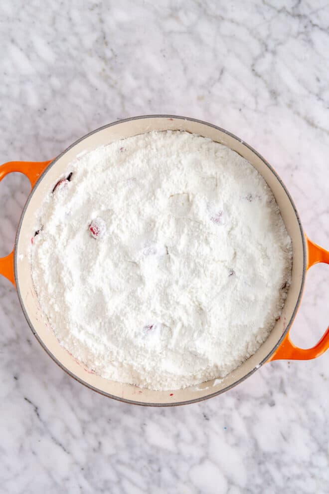 Frozen berries topped with yellow cake mix in a white enamel lined dutch oven with orange handles.