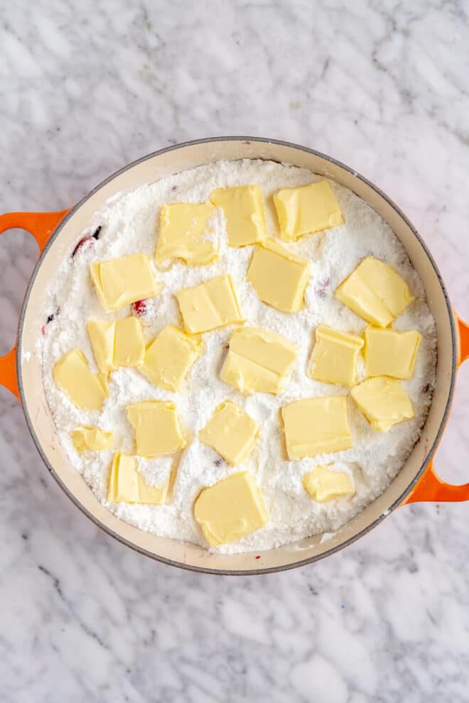 Pads of butter topping yellow cake mix in a dutch oven with orange handles on a grey and white marble surface.