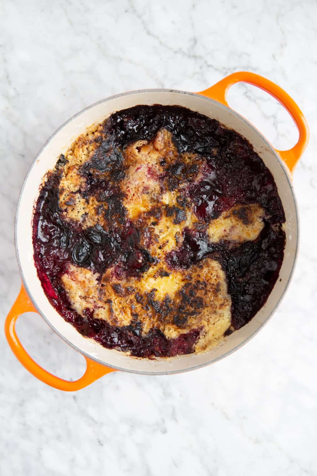 Baked triple berry dump cake in a dutch oven with orange handles on a grey and white marble surface.