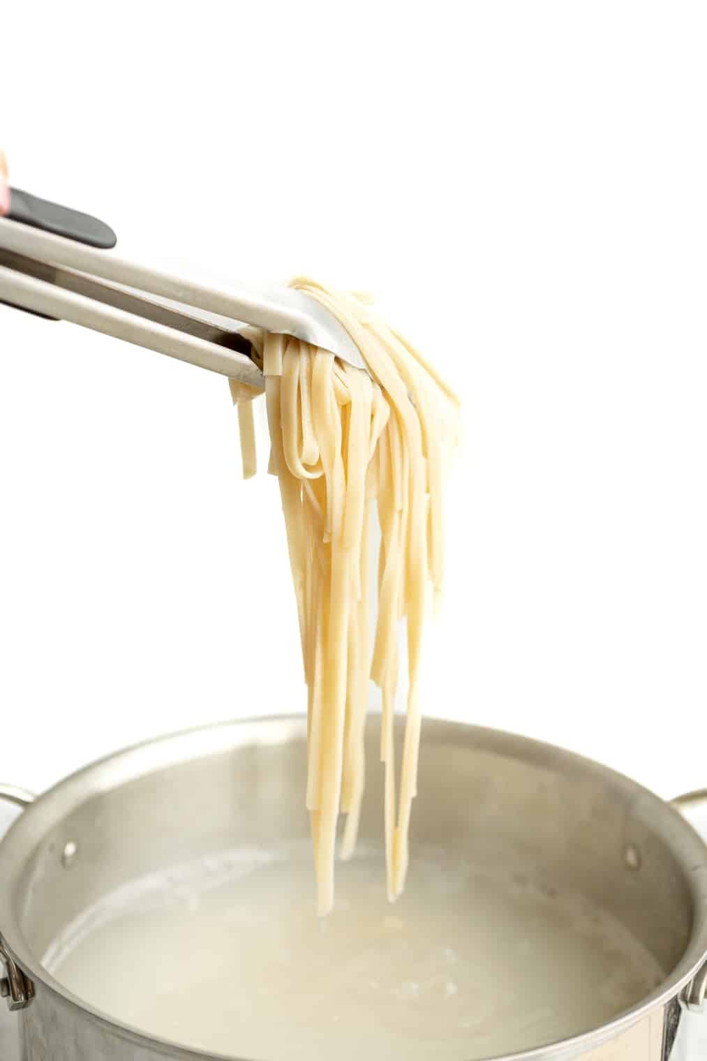 A person using tongs to hold al dente fettuccine noodles above a pot of pasta water.