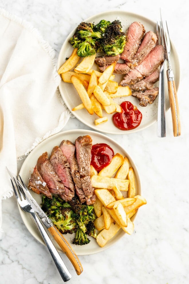 Two dinner plates with sliced steak, broccoli, and steak fries with a side of ketchup.