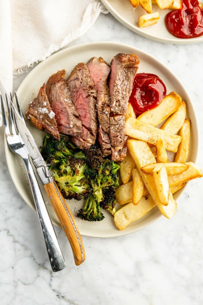 Sliced steak plated with broccoli and steak fries with a side of ketchup.