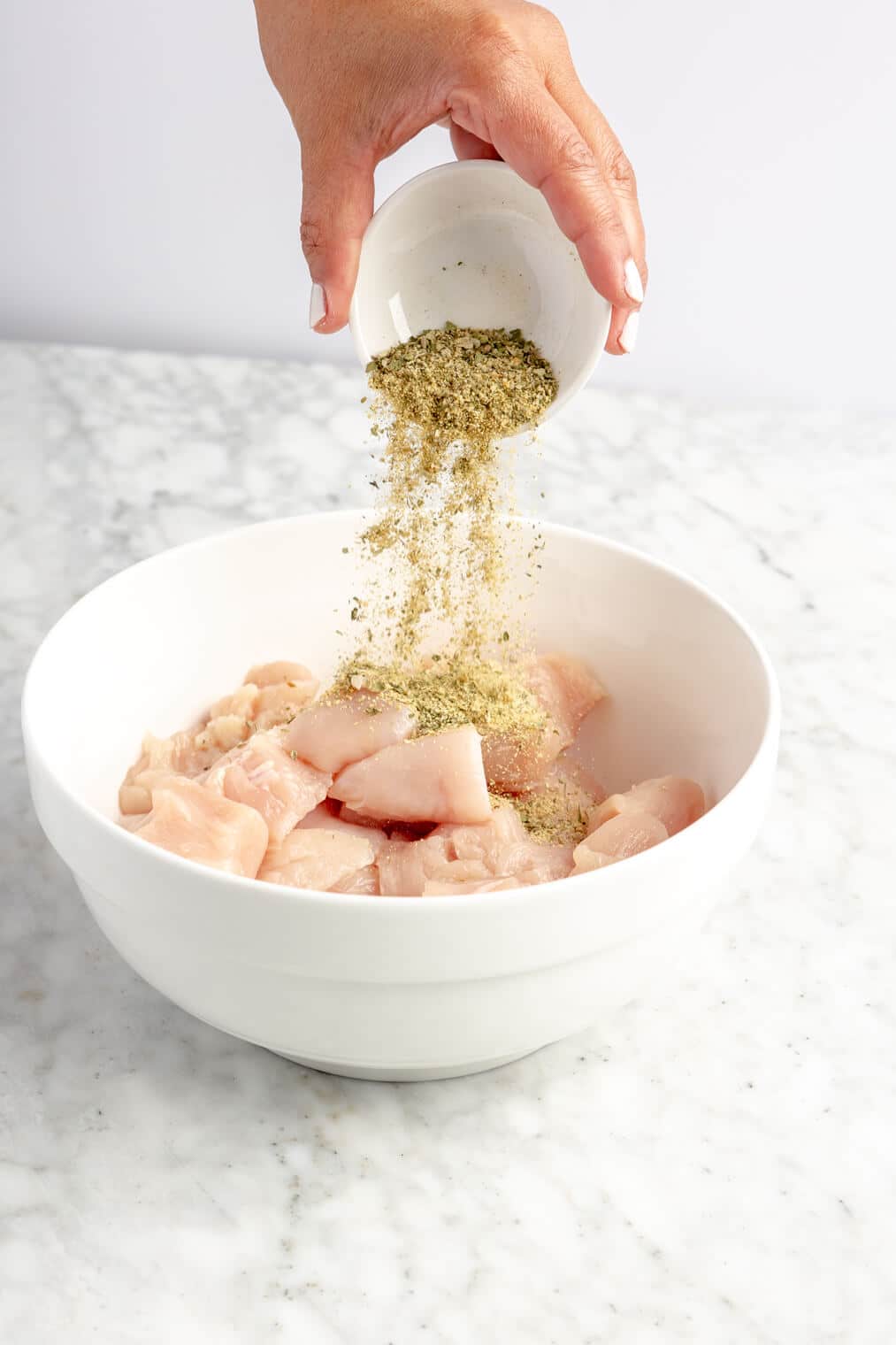 Hand holding small, white bowl with seasoning sprinkling over a white bowl with cubed chicken breast.