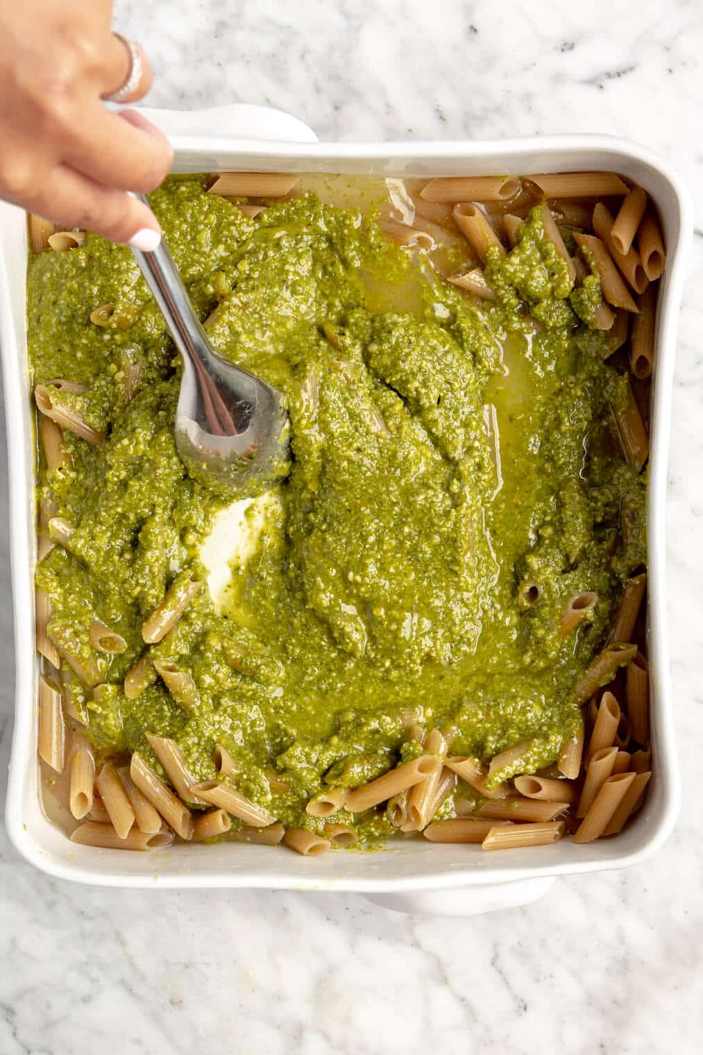 Hand stirring pesto and broth in casserole dish with noodles.