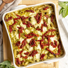 Top down view of white, square casserole dish with pesto pasta topped with sun-dried tomatoes and mozzarella on a wooden cutting board.