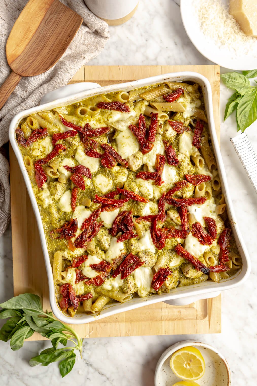 Top down view of white, square casserole dish with pesto pasta topped with sun-dried tomatoes and mozzarella on a wooden cutting board.