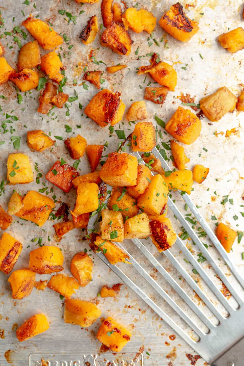 Roasted butternut squash garnished with fresh herbs on a sheet pan with a spatula serving some of the squash.