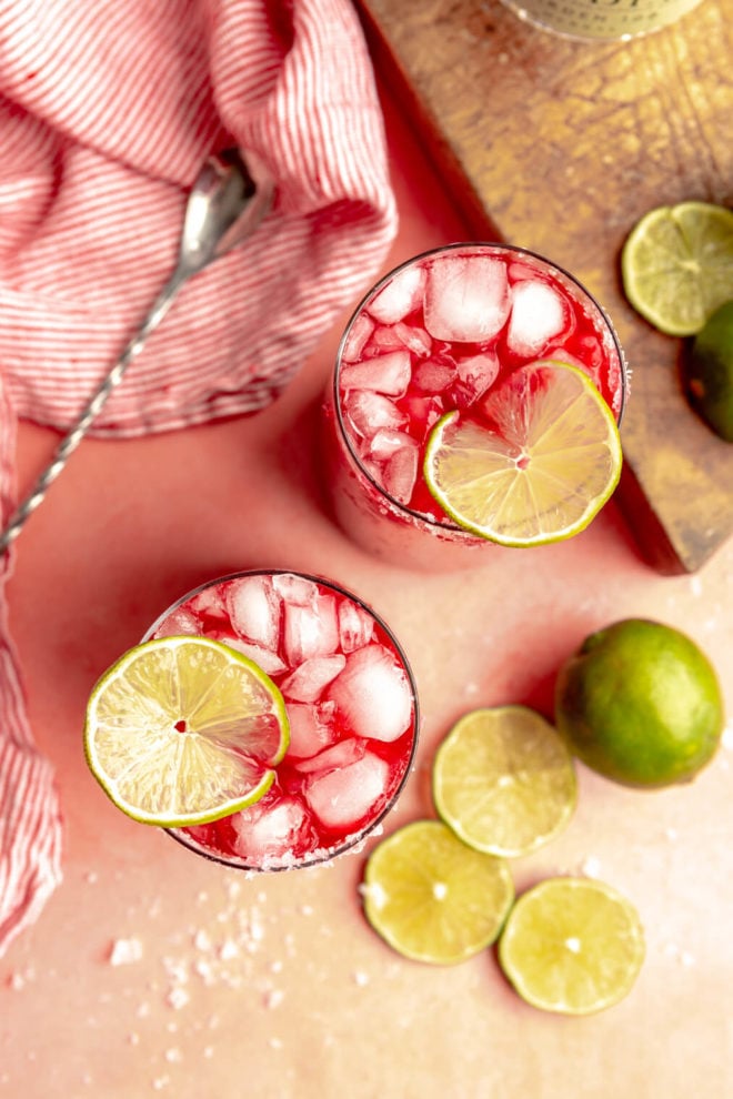 Top down of two glasses with red drink over ice. They are garnished with thin, round slices of lime and sitting on a pink table. There is a red and white striped linen and limes on the table.