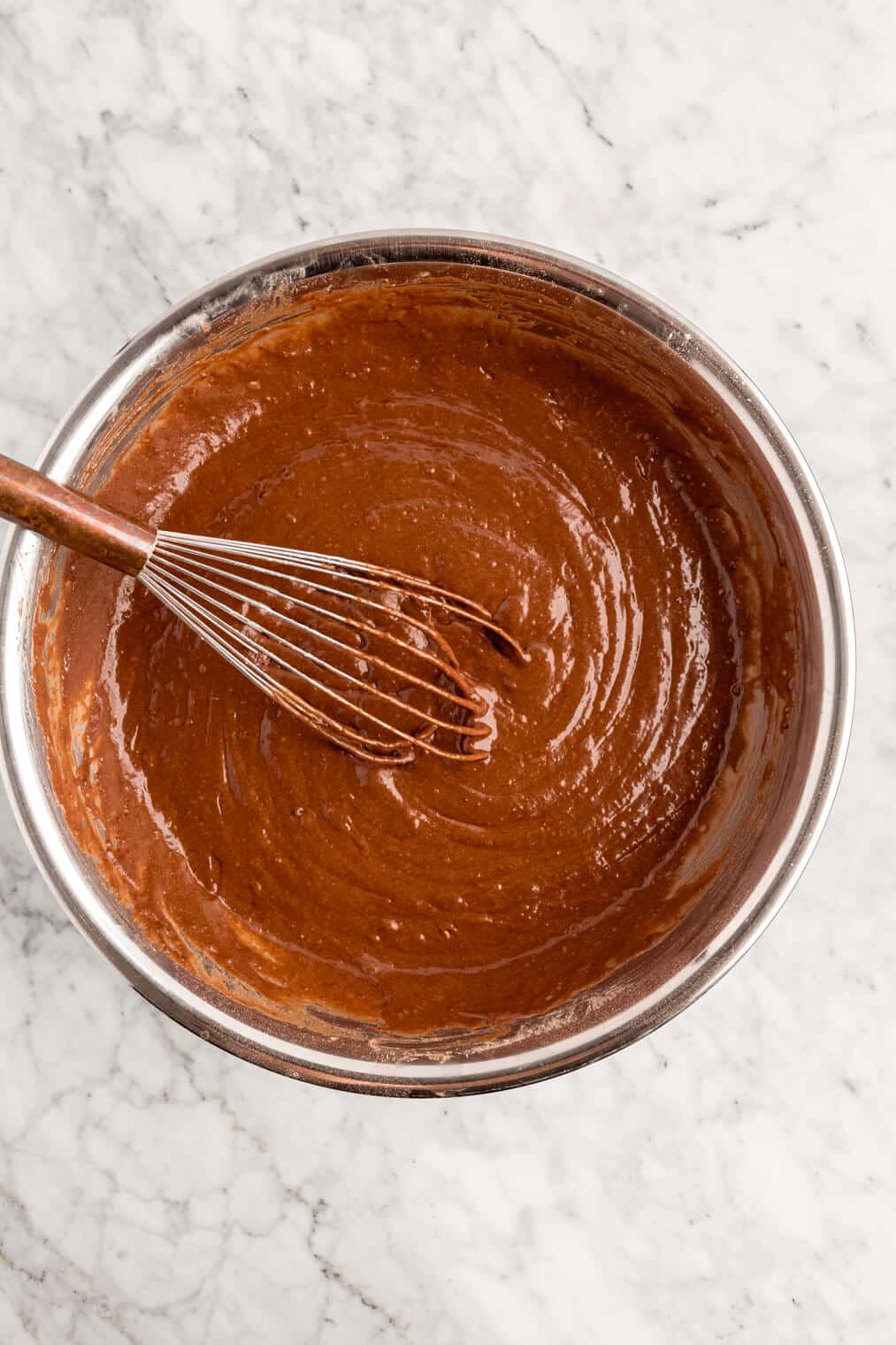 Chocolate cake batter whisked until smooth in a large metal bowl on a grey and white marble surface.
