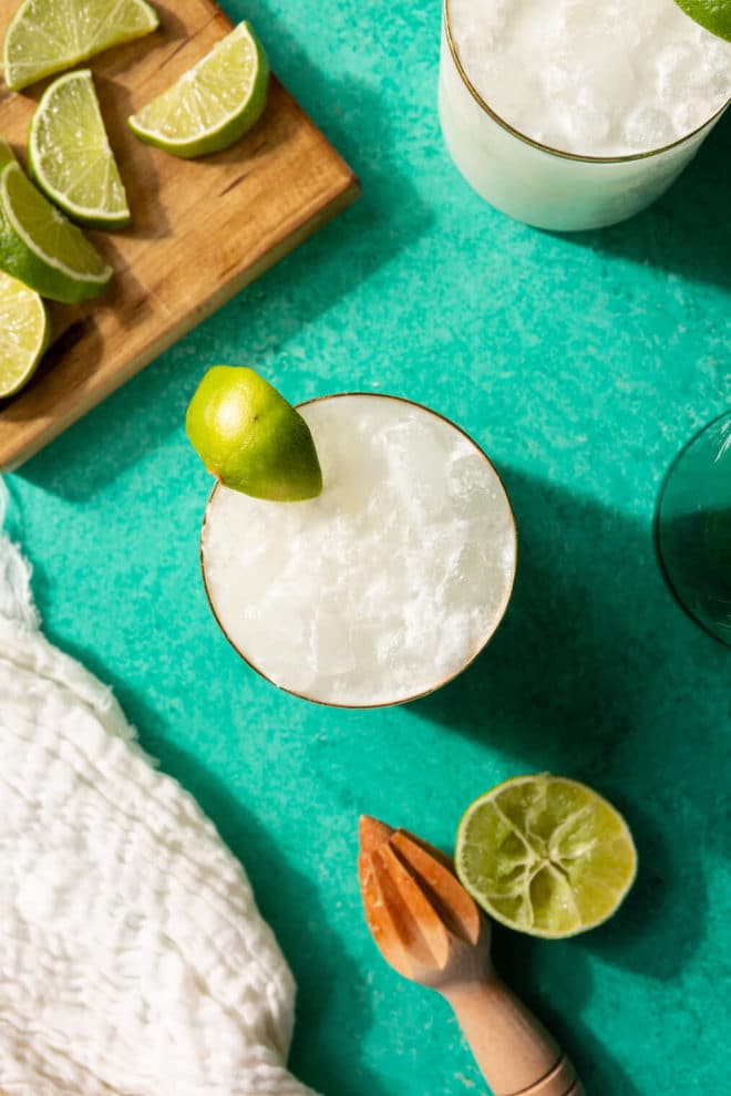 Top down of a gold rimmed glass with a creamy white beverage on ice inside. There is also a wooden cutting board with lime wedges and a lime half with a citrus pestle. All are on a turquoise surface.
