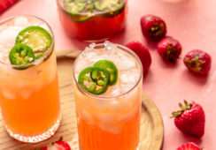 Two tall glasses with light pink/red liquid and ice garnished with sliced jalapeños on a wound, wooden cutting board on a pink surface with sliced strawberries, jalapeños, and a mason jar with more mocktail drink inside and a bottle of Seedlip spirits in the background.