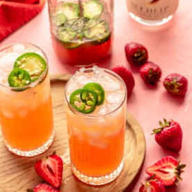 Two tall glasses with light pink/red liquid and ice garnished with sliced jalapeños on a wound, wooden cutting board on a pink surface with sliced strawberries, jalapeños, and a mason jar with more mocktail drink inside and a bottle of Seedlip spirits in the background.