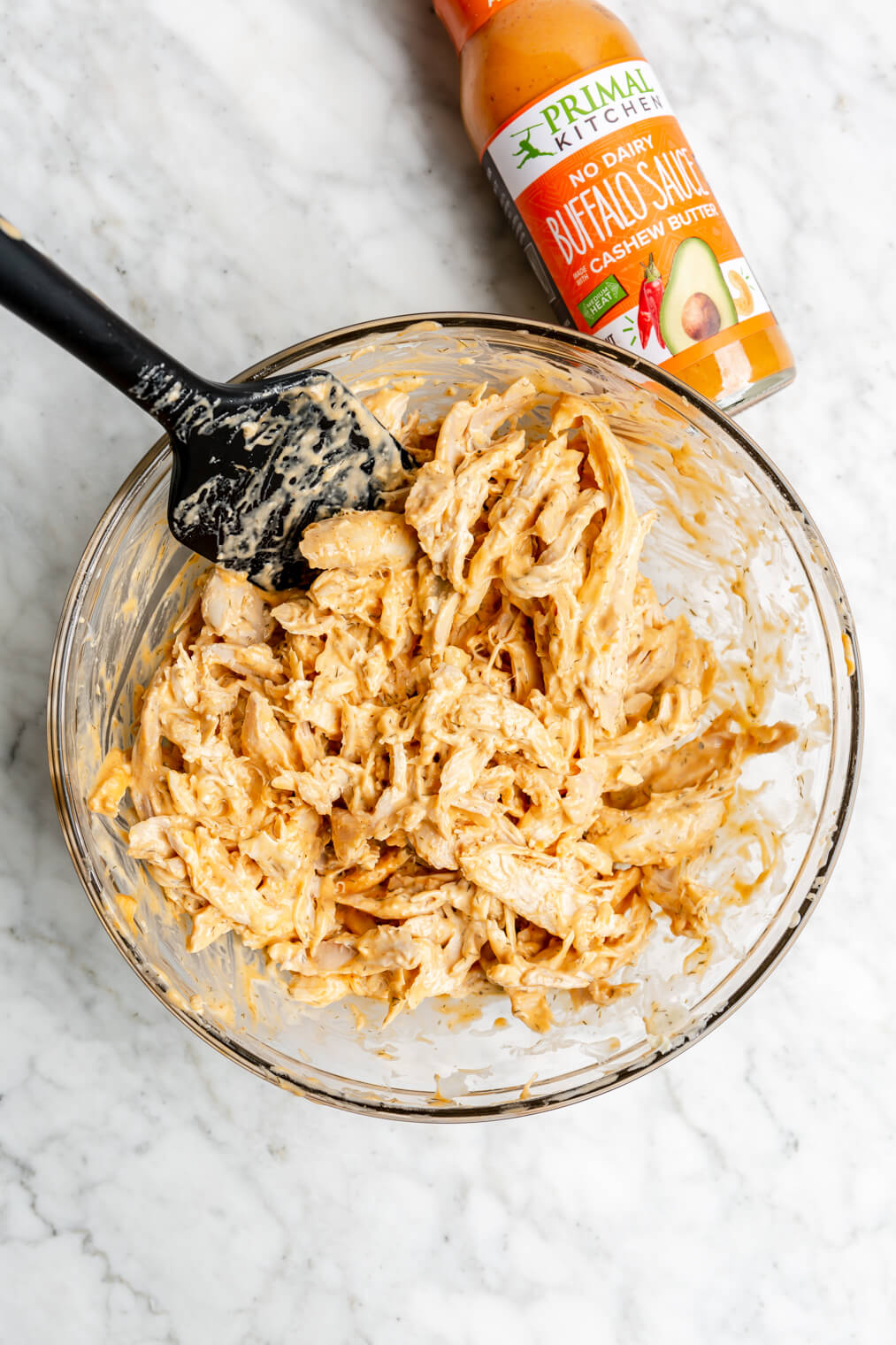 Shredded chicken in a buffalo and ranch mixture in a glass bowl on a gray and white surface.