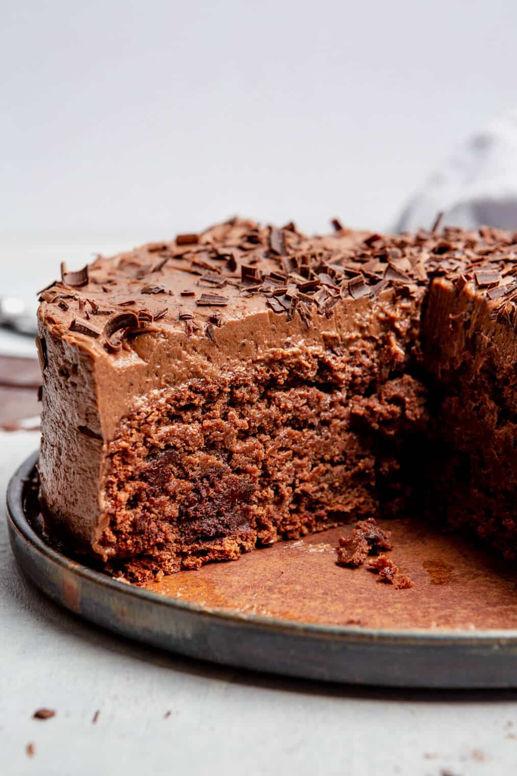 Side view of a single layer chocolate cake coated with chocolate frosting and shaved chocolate on top. The cake is sitting on a black plate with a rust colored brown interior and has a slice removed.
