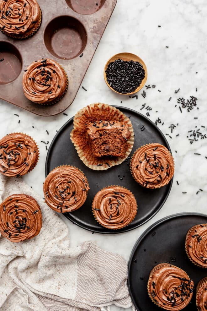 Top down view of chocolate cupcakes on a black plate with some cupcakes on the counter to the side and come in the cupcake tin with a bowl of chocolate sprinkles and chocolate sprinkles scattered on the surface.
