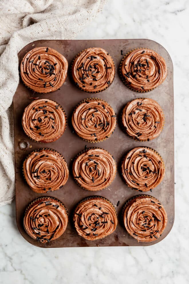 Top down view of a brown muffin tin with 12 chocolate cupcakes topped with chocolate frosting and chocolate sprinkles. The muffin tin is sitting on a grey and white marble surface and has a linen draped to the top left corner.