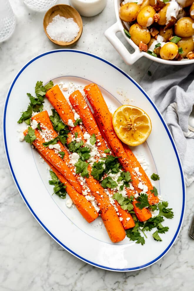 White oval serving dish with large carrots garnished with cilantro and goat cheese. There is a lemon half on the side that has been squeezed.