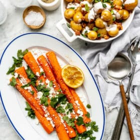 Full carrots plated on a white oval serving dish with a blue rim garnished with cilantro and goat cheese crumbles and a squeeze lemon half. Next to that dish is a bowl of roasted Yukon gold potatoes drizzled with a cream and topped with bacon and herbs. A small bowl of flaky sea salt and ground pepper are also on the table along with serving utensils.