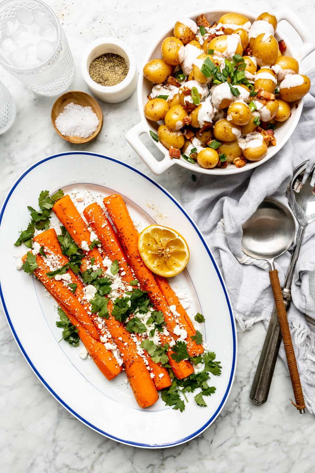 Full carrots plated on a white oval serving dish with a blue rim garnished with cilantro and goat cheese crumbles and a squeeze lemon half. Next to that dish is a bowl of roasted Yukon gold potatoes drizzled with a cream and topped with bacon and herbs. A small bowl of flaky sea salt and ground pepper are also on the table along with serving utensils.