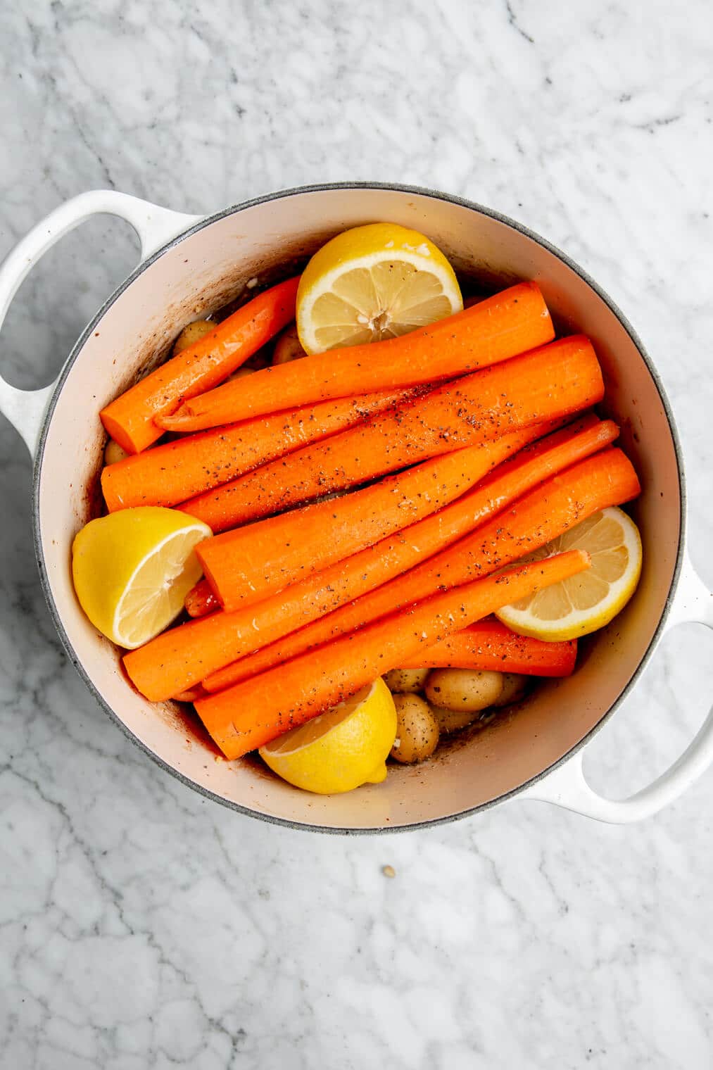 Full carrots layered over Yukon gold potatoes with lemon halves nestled alongside in a large, white dutch oven.