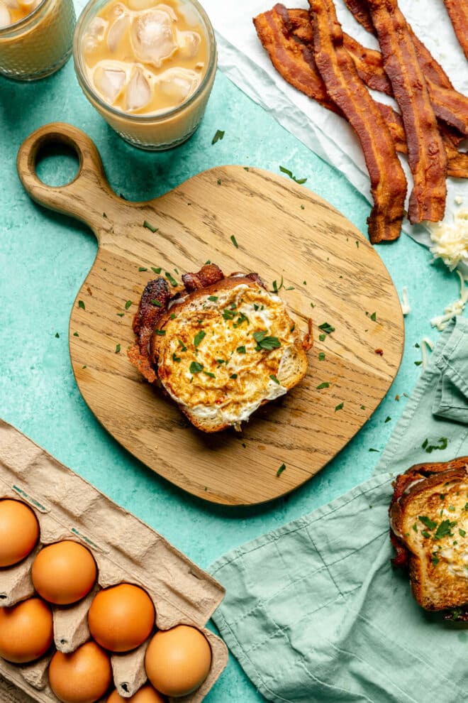 Egg-in-a-hole grilled cheese sandwich on a wooden cutting board with a carton of eggs, pile of bacon, cup of iced coffee in the background on a teal surface. 