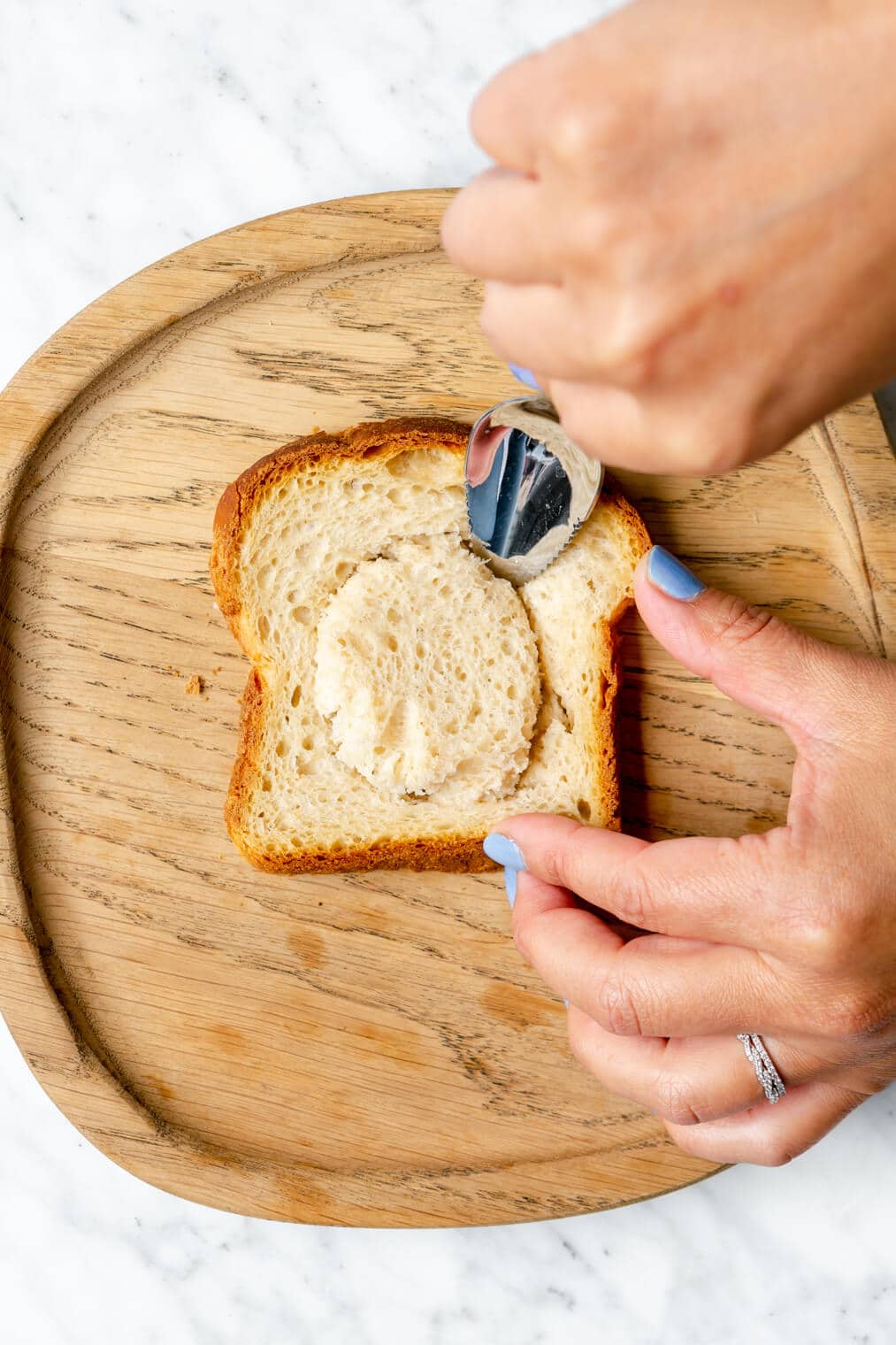 Hand holding a spoon cutting out a circle in a slice of bread on a wooden cutting board.