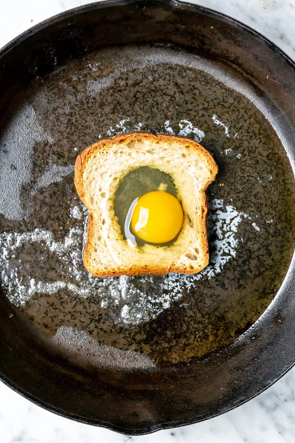Slice of bread with hole in middle with an egg cracked in the hole in a cast iron skillet with melted butter.